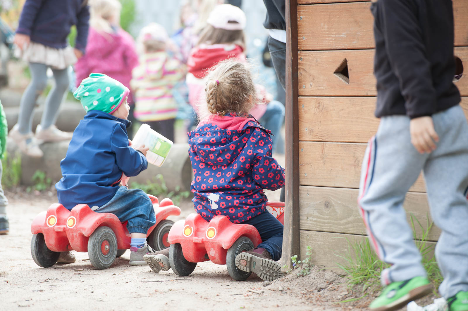 Zwei Kinder von hinten auf Bobbycars vor dem Holz-Spielhaus