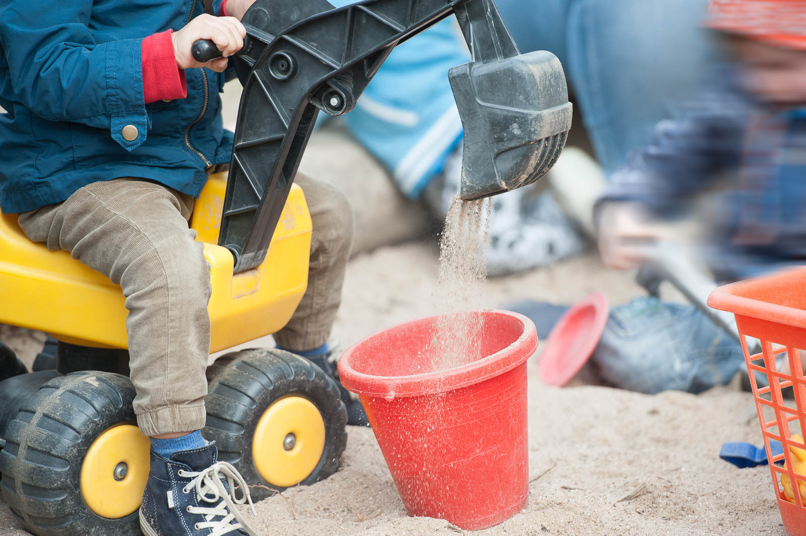 Kind spielt im Sandkasten des Gartens und schüttet Sand mit einem Spielzeugbagger in einen roten Eimer